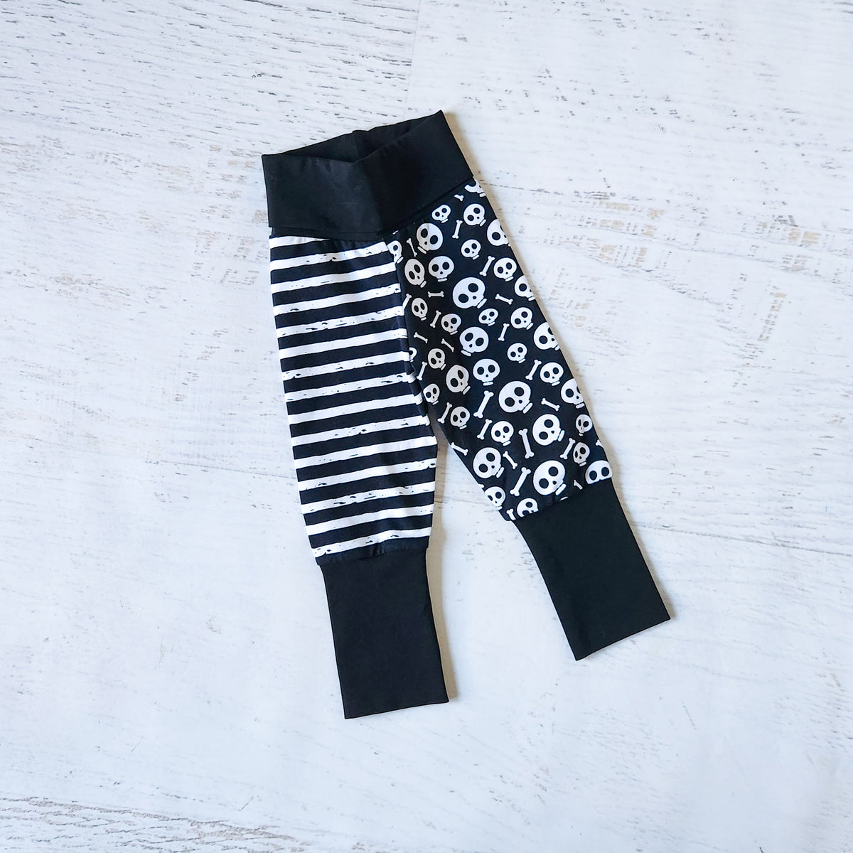 Black and white patterned baby jogger pants laid flat on a white distressed wood background. The pants feature a black and white striped leg, a black leg with a white skull print, and solid black cuffs.