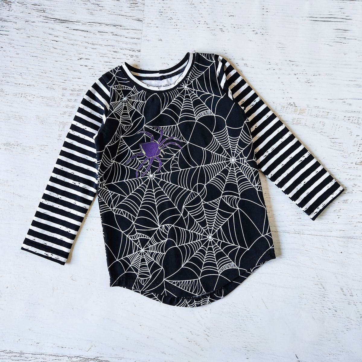 A children's long-sleeve top laid flat on a white rustic wood background. The shirt has black-and-white striped sleeves, a black body with a white spiderweb print, and a purple spider applique on the chest.